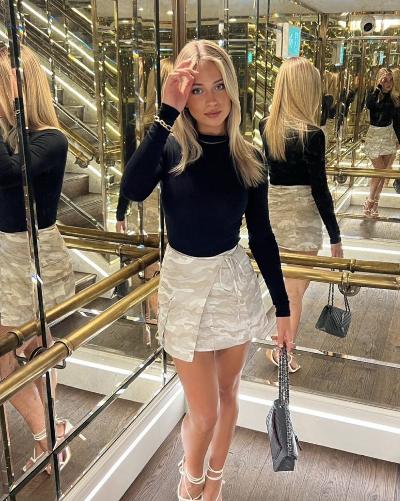 Woman in an elegant mirrored elevator wearing a black turtleneck, patterned skirt, and white strappy heels, accessorized with a chic handbag.